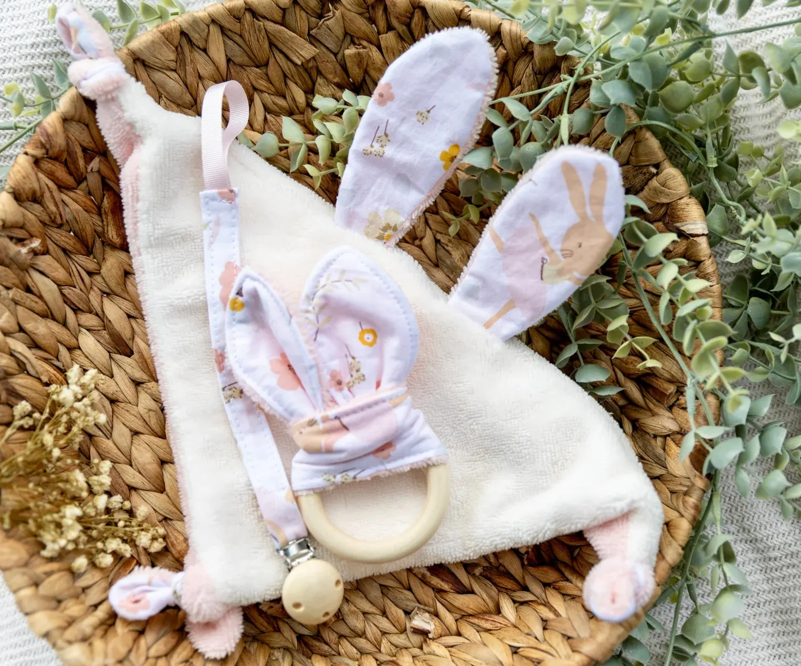 Ensemble doudou plat lapin écru et rose, attache-tétine et anneau de dentition dans une corbeille tressée