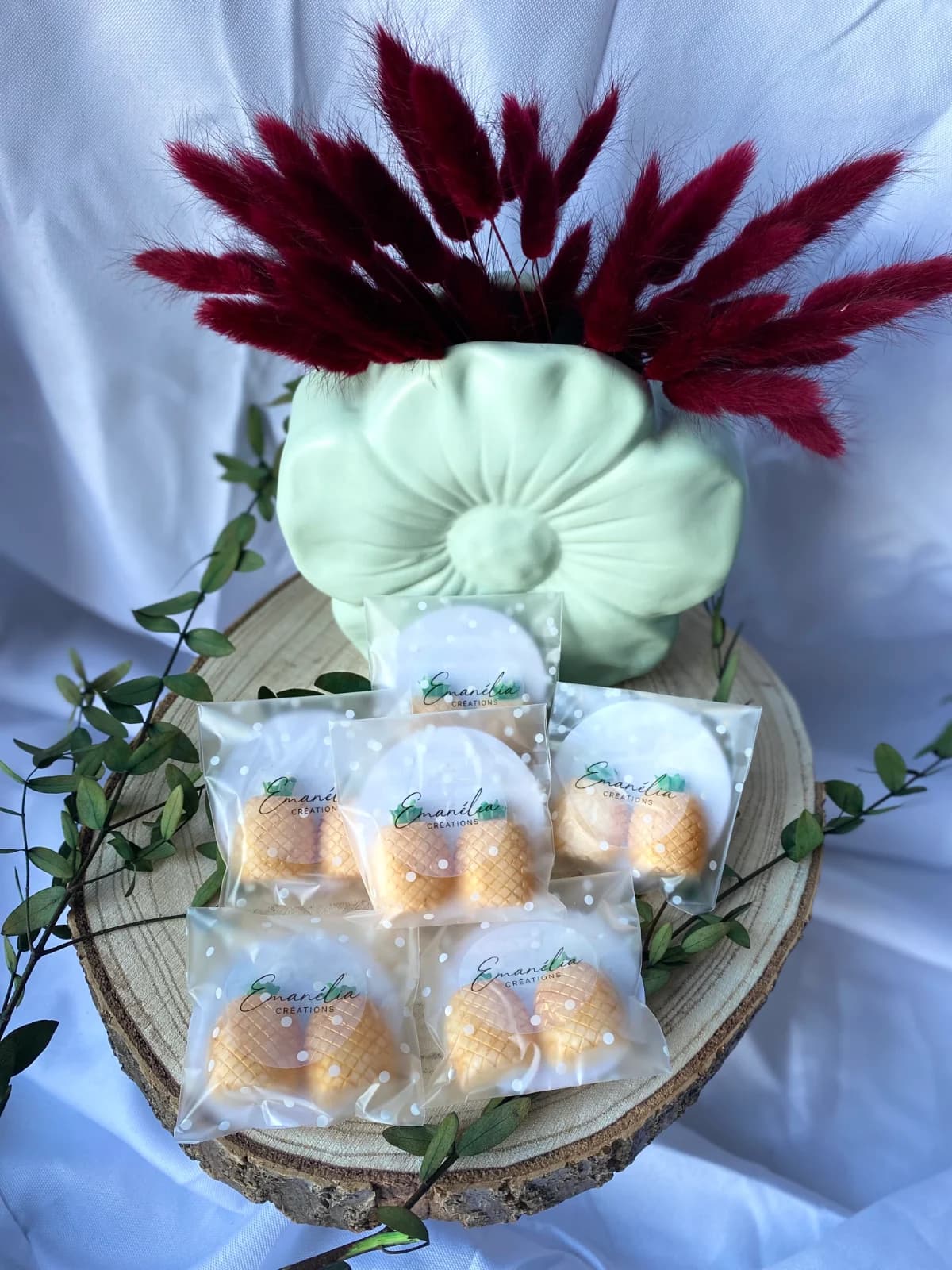 Vue d'ensemble des sachets de fondants ananas sur tranche de bois avec vase vert et fleurs séchées bordeaux.